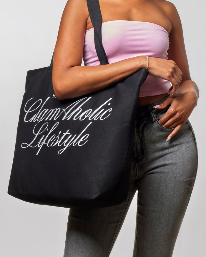 Black canvas tote bag featuring "Glam-Aholic Lifestyle" logo, styled with a model in casual attire.