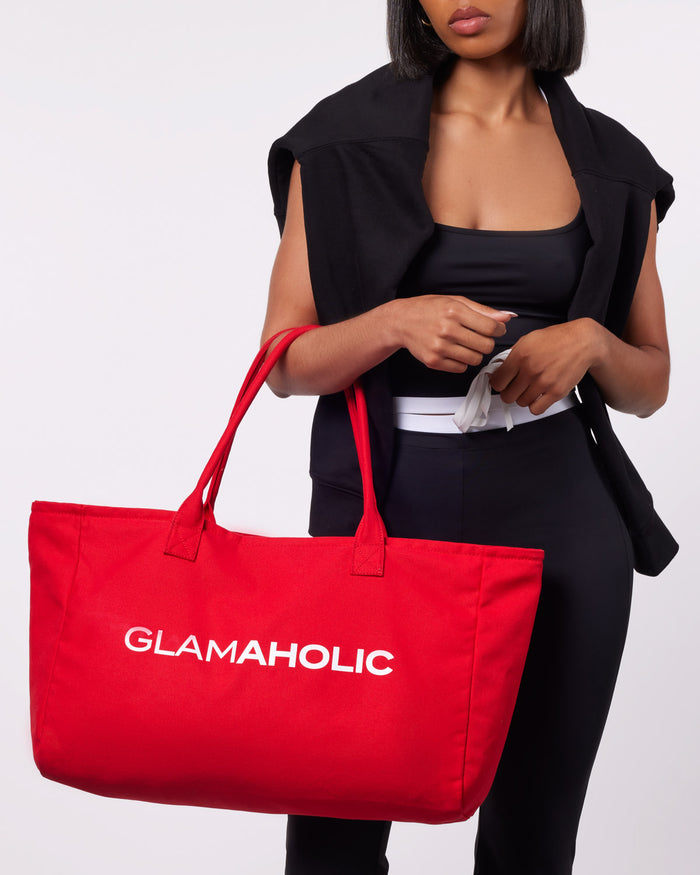 Red athletic canvas tote bag with "GLAMAHOLIC" logo, held by a model in a black outfit. Ideal for stylish storage.