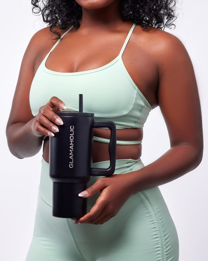 A woman in a mint green athletic outfit holds a black athletic tumbler with a straw, featuring the Glam-Aholic logo.
