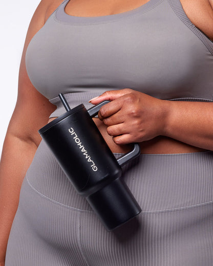 A person holds a black athletic tumbler with a straw, wearing a gray sports bra and matching leggings.