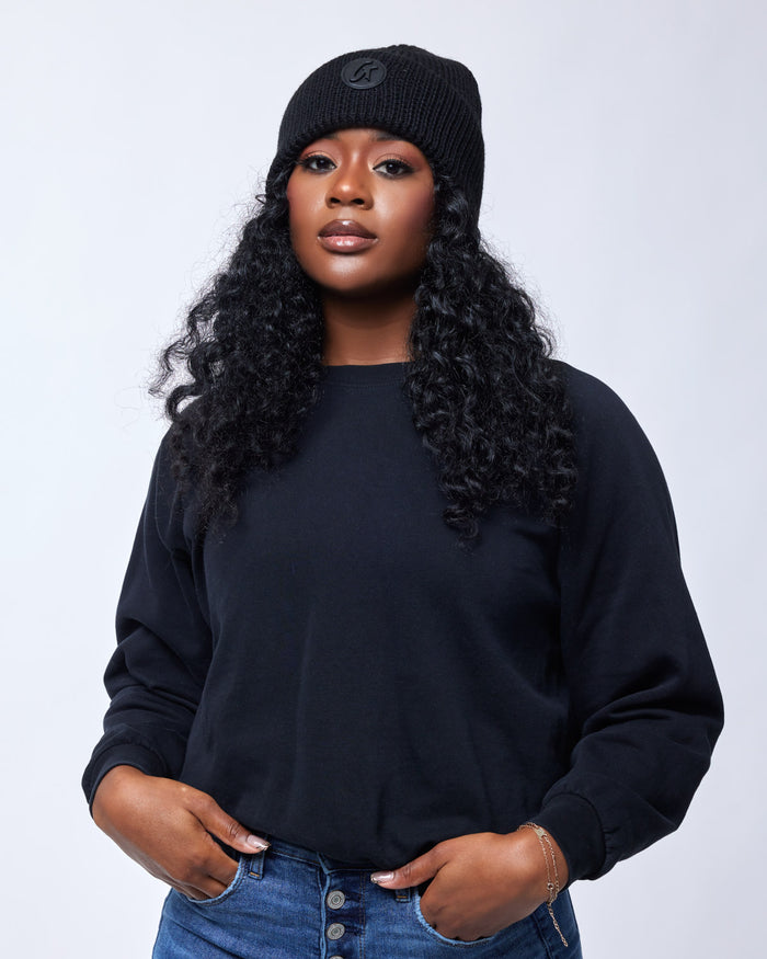 Black classic beanie hat with a logo patch, worn by a woman with curly hair, black sweatshirt, and blue jeans.