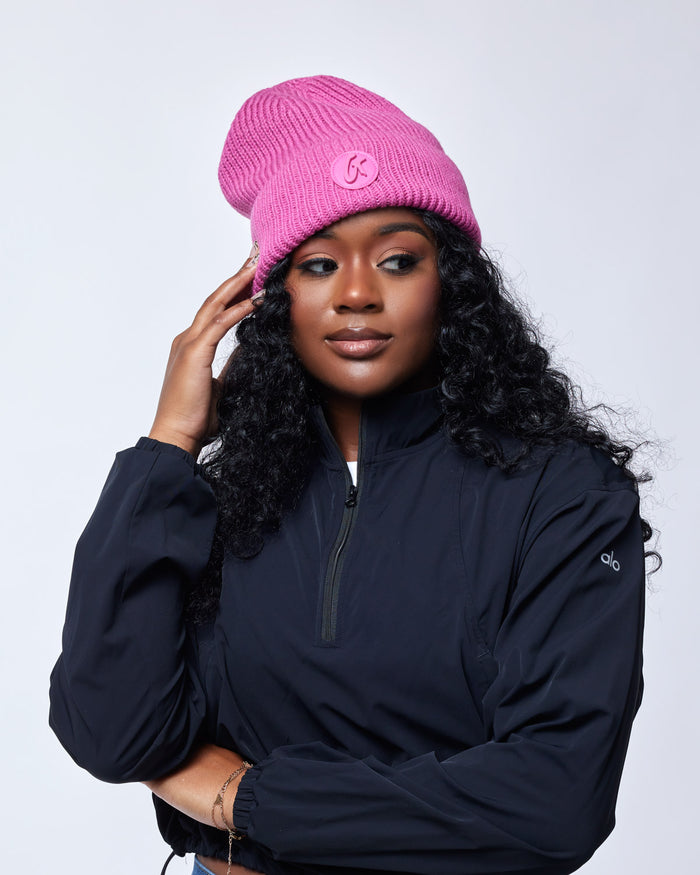 Woman wearing a bright pink classic beanie with a logo, dressed in a black zip-up jacket, against a plain background.