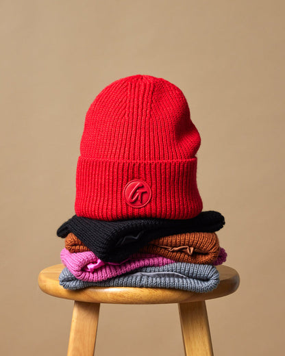 Red classic beanie displayed on top of a stack of colorful knit hats on a wooden stool.