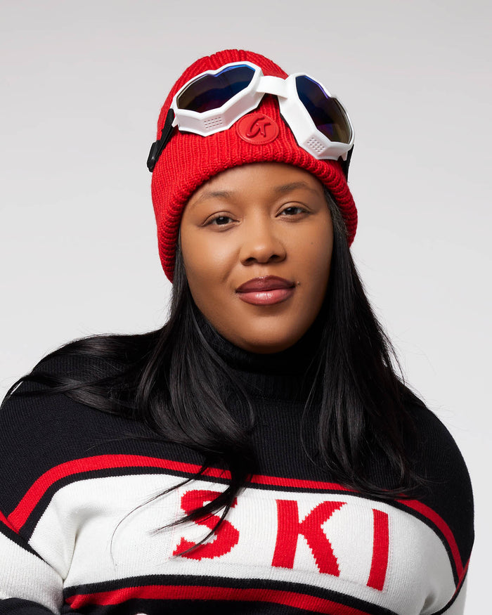 Woman wearing a red knit beanie with goggles on top, black sweater with red and white "SKI" lettering.