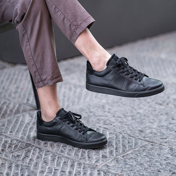 Black leather sneakers with lace-up closure on textured pavement with pants visible.