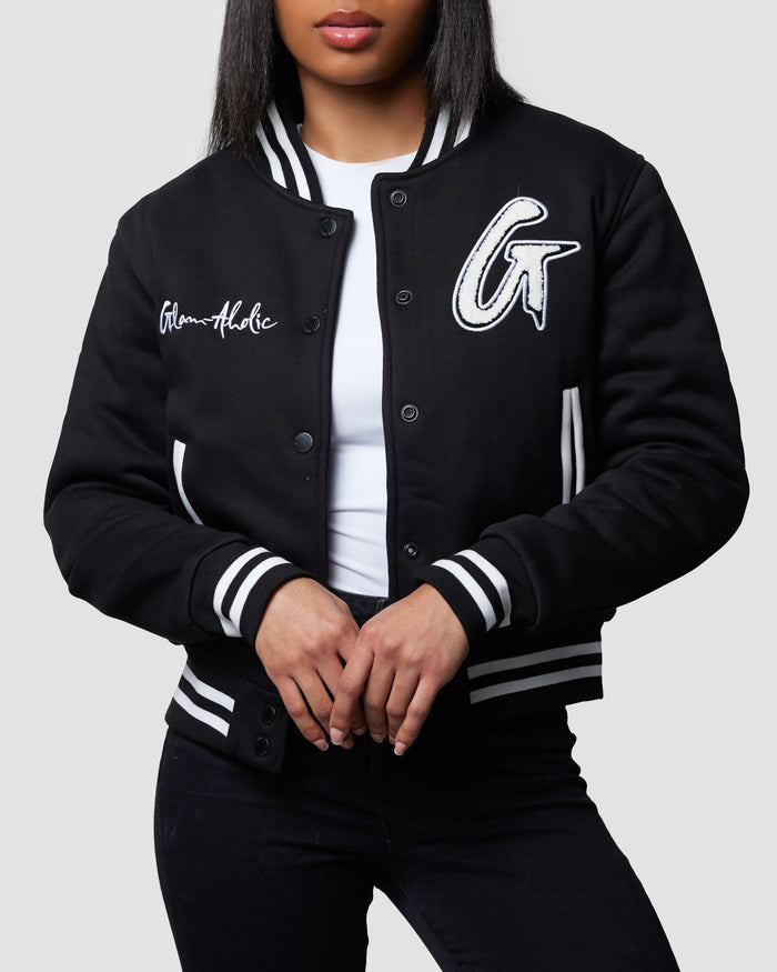 Black varsity jacket with white striped cuffs, collar, and hem, featuring embroidered patches and text, worn over a white top.