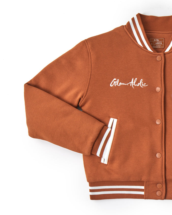 Brown varsity jacket with striped cuffs, embroidered "Glam-Aholic" logo, snap buttons, and ribbed collar and hem.