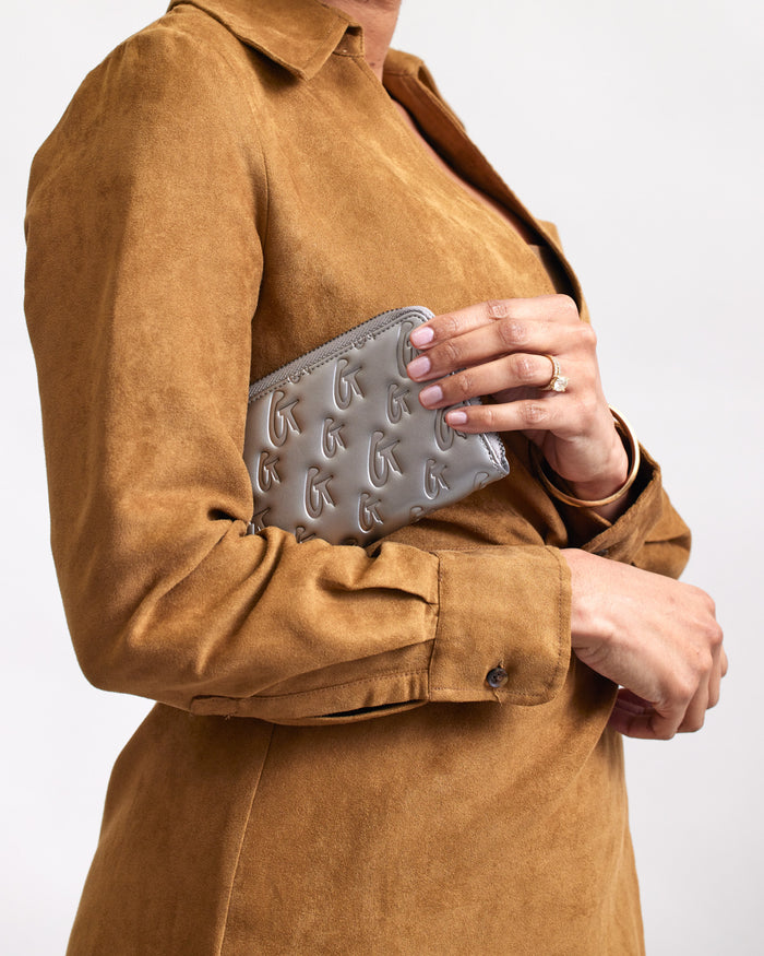 A person in a brown suede jacket holds a gray jumbo monogram wallet with embossed logos, clutching it against their chest.