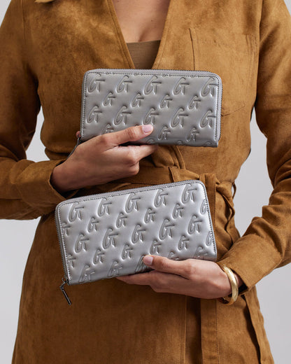 Two silver jumbo monogram wallets with embossed "G" pattern, held by a person in a brown suede coat.