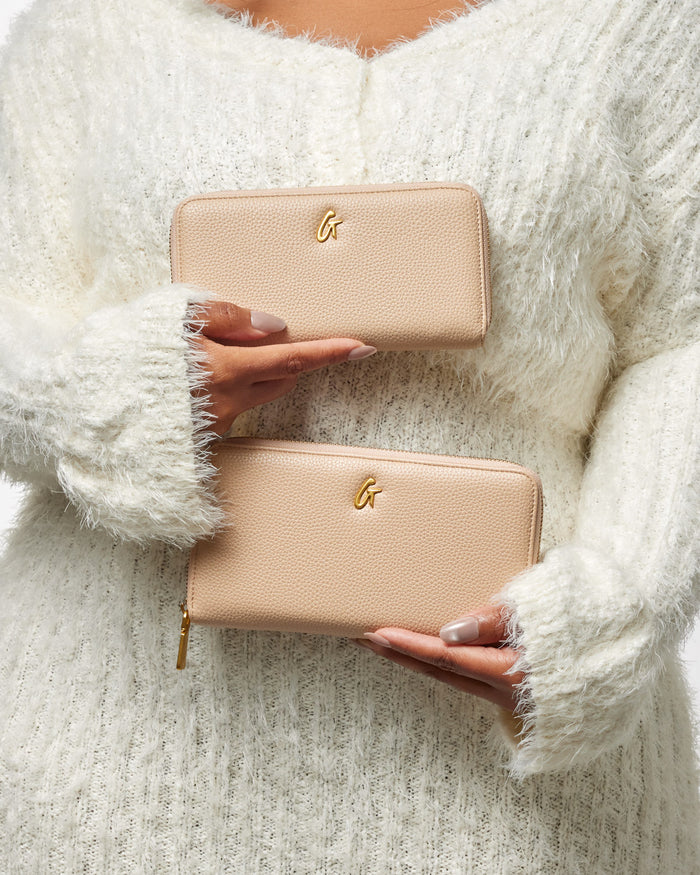 Two beige pebble-textured wallets with gold "G" logos, held by a person in a cozy white sweater.