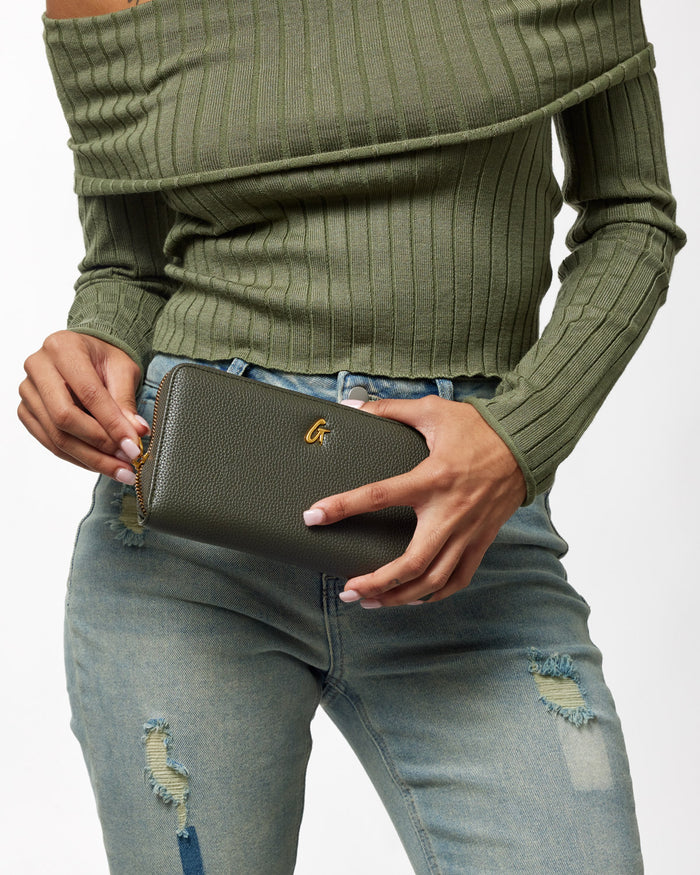 Olive green jumbo pebble leather wallet with gold hardware, held by a person wearing a green ribbed sweater and distressed jeans.