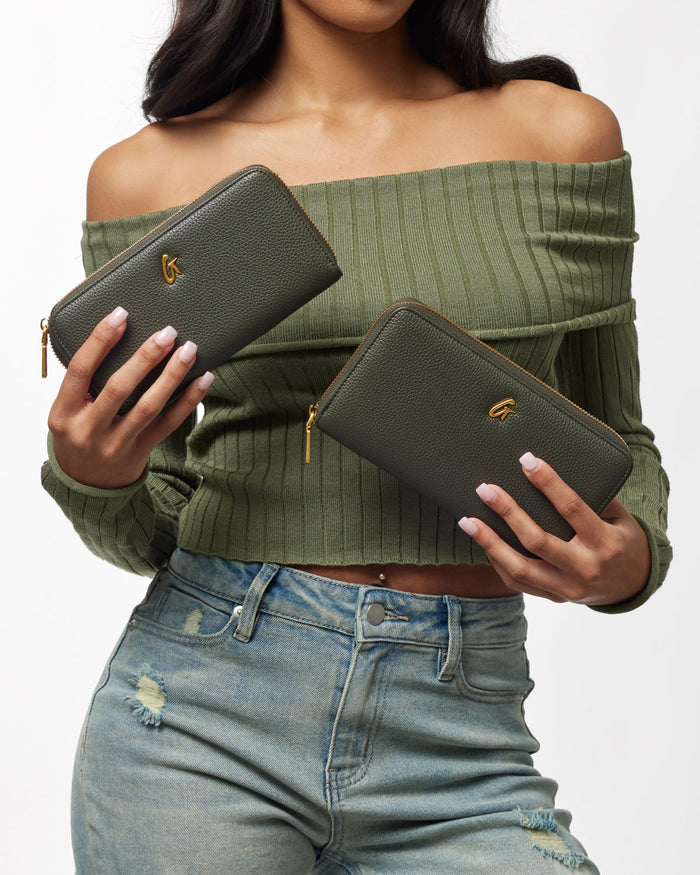 Olive green jumbo pebble wallets with gold zipper and logo, held by a woman wearing a green off-shoulder top and distressed jeans.