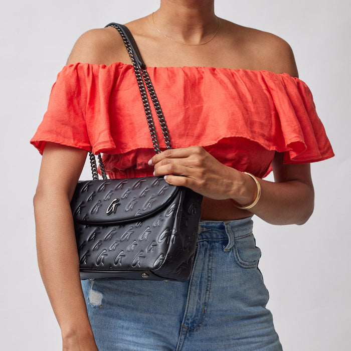 Black medium monogram flap bag with embossed logo pattern, worn over a red off-shoulder top and blue jeans.
