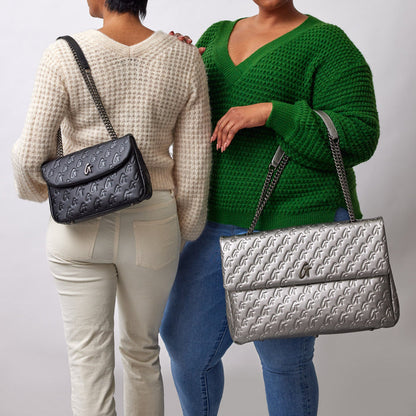 Two women hold black and silver medium monogram flap bags with chain straps, featuring a repeating logo pattern.
