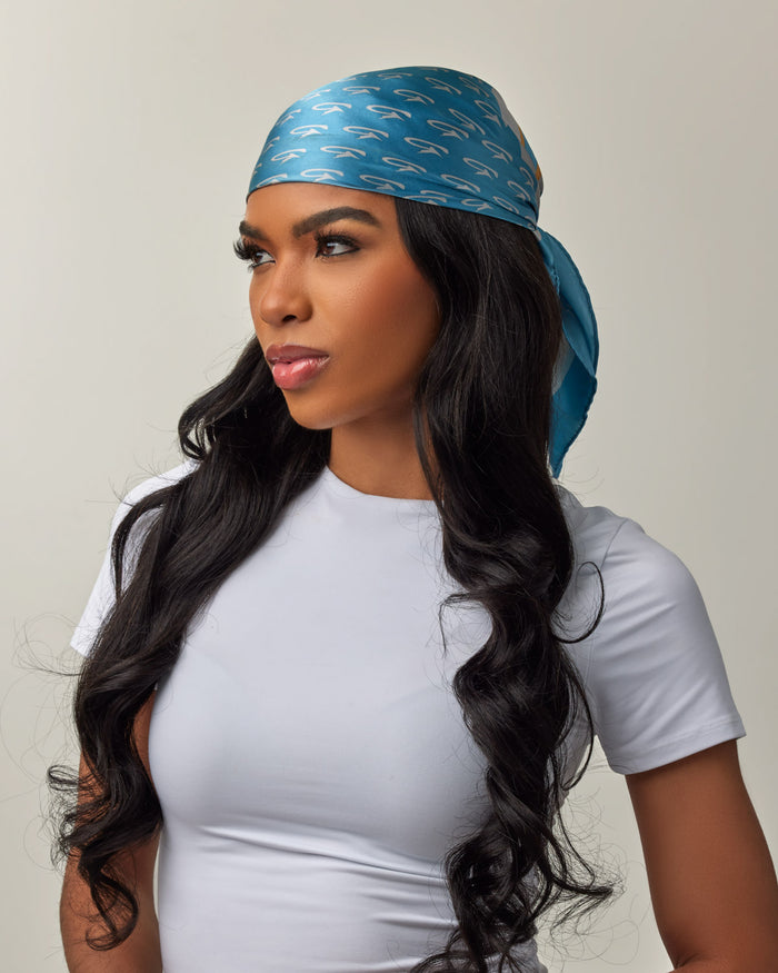 A woman wears a blue silk scarf with gold monogram pattern, styled as a head wrap, paired with a white t-shirt.