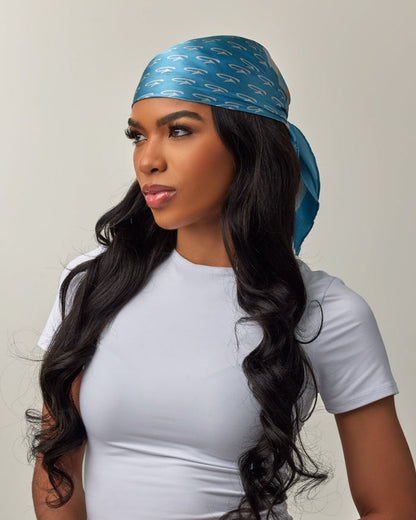 A woman wears a blue silk scarf with gold monogram pattern, styled as a head wrap, paired with a white t-shirt.