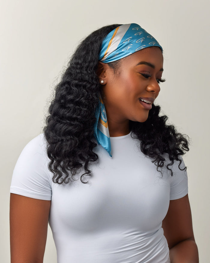 A woman wears a blue and gold silk scarf with monogram pattern, styled as a headscarf, against a plain light background.