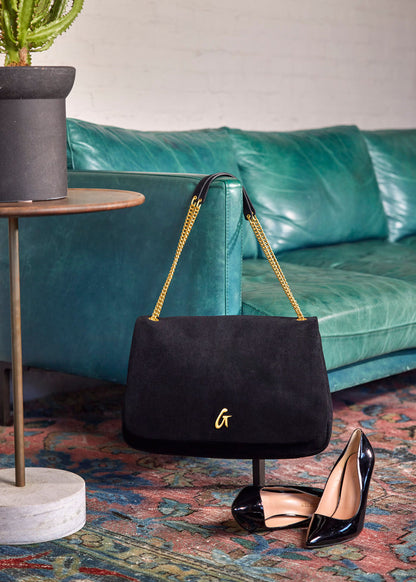 Medium black suede flap bag with gold chain strap displayed beside black high heels on a colorful rug.