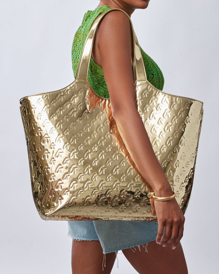 Woman holding a large metallic gold tote bag with embossed monogram pattern, paired with a green crochet top and denim shorts.