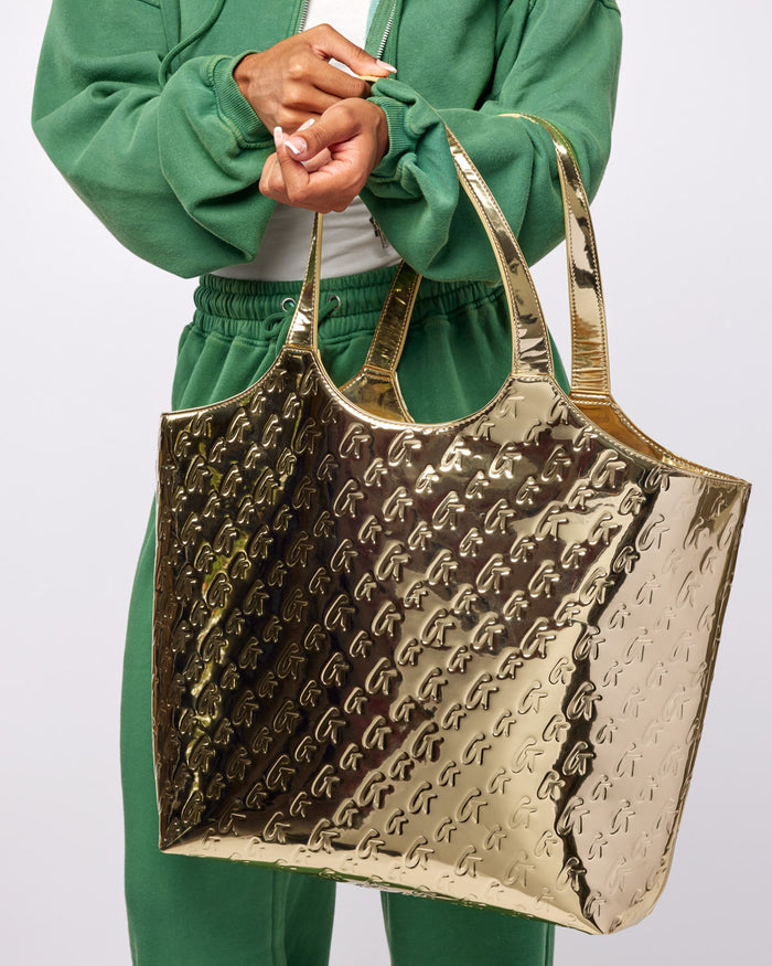 Shiny metallic gold maxi tote with a monogram design, held by a person in a green outfit.