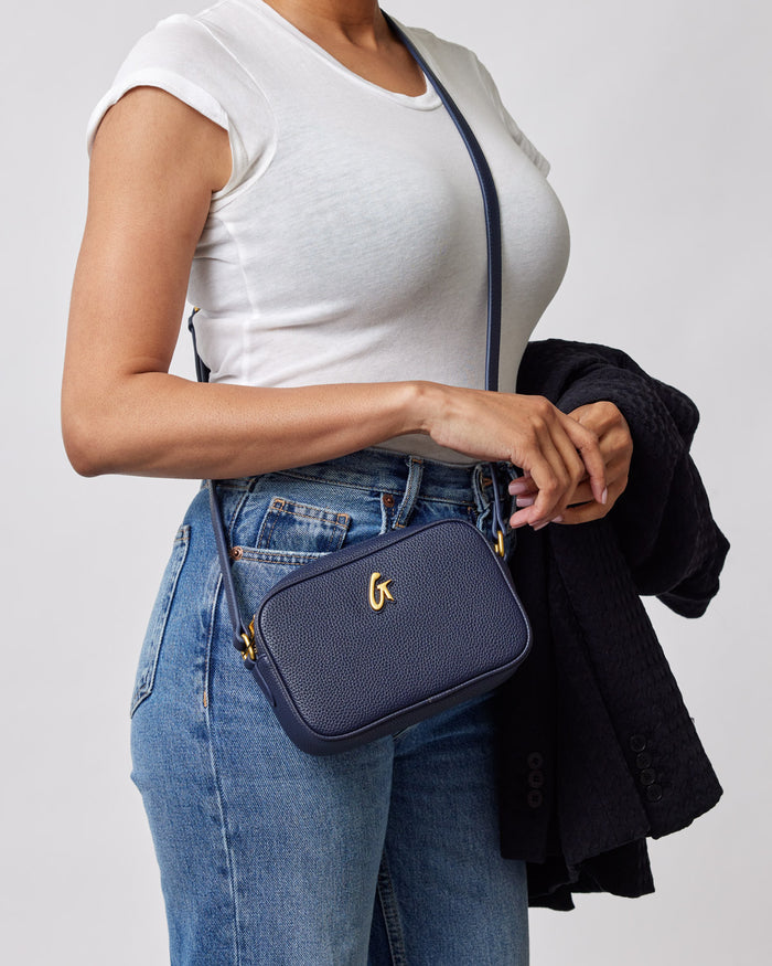 Mini Pebble Camera Bag in navy, with a textured surface, gold logo, and adjustable strap, worn crossbody by a woman.