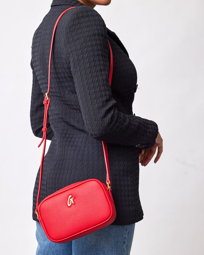Red mini pebble camera bag with a gold logo, worn crossbody over a black textured blazer.