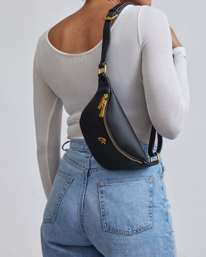 Black Pebble Fanny Pack with gold zipper and hardware, worn crossbody on a woman in a white long-sleeve top and blue jeans.