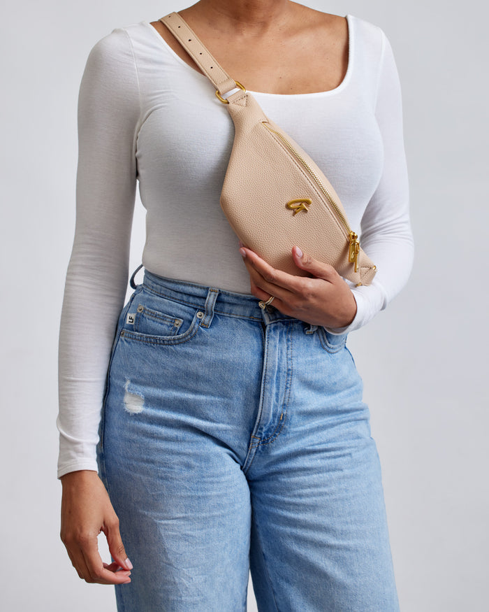 Mini pebble nude fanny pack worn across the chest, featuring a gold zipper and logo, paired with a white long-sleeve top and high-waisted blue jeans.