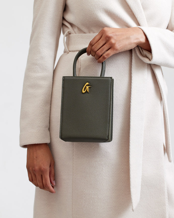 Mini pebble leather tote in olive green with a gold logo, held by a person wearing a cream-colored coat.