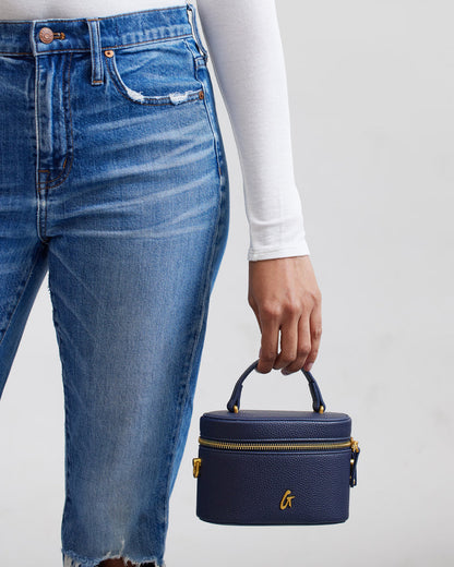 Mini navy pebble vanity bag with gold zipper and handle, held by a person wearing blue jeans and a white long-sleeve shirt.