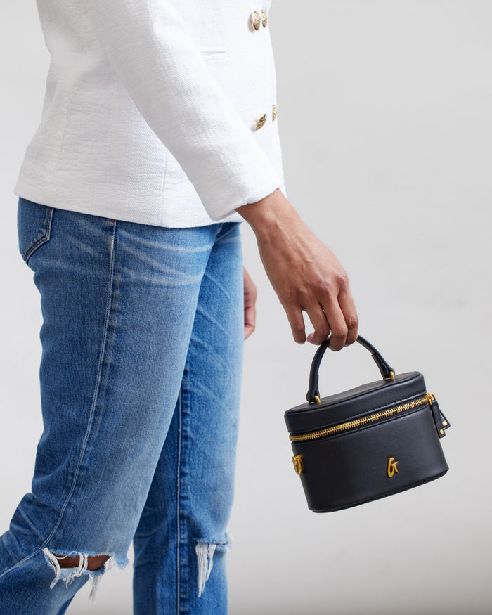 Mini black vanity bag with gold accents, top handle, and zipper closure, held by a person wearing a white blazer and ripped jeans.