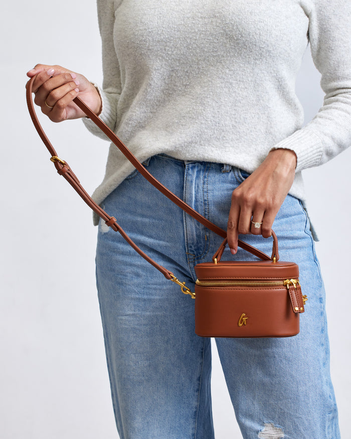 Mini smooth brown vanity bag with gold accents, a top handle, and adjustable strap, held by a person in casual clothing.