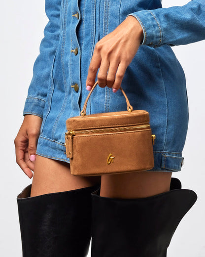 Brown mini suede vanity bag with gold zipper, held by a model wearing a denim dress and knee-high black boots.