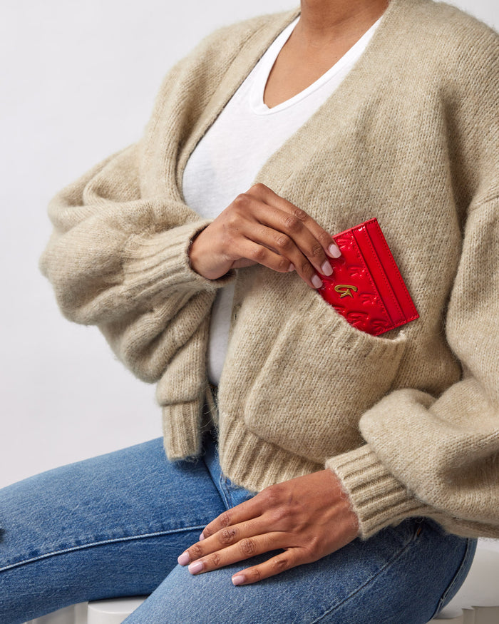 Red mirror-like card holder with gold monogram, partially tucked into the chest pocket of a beige knit cardigan worn by a person.