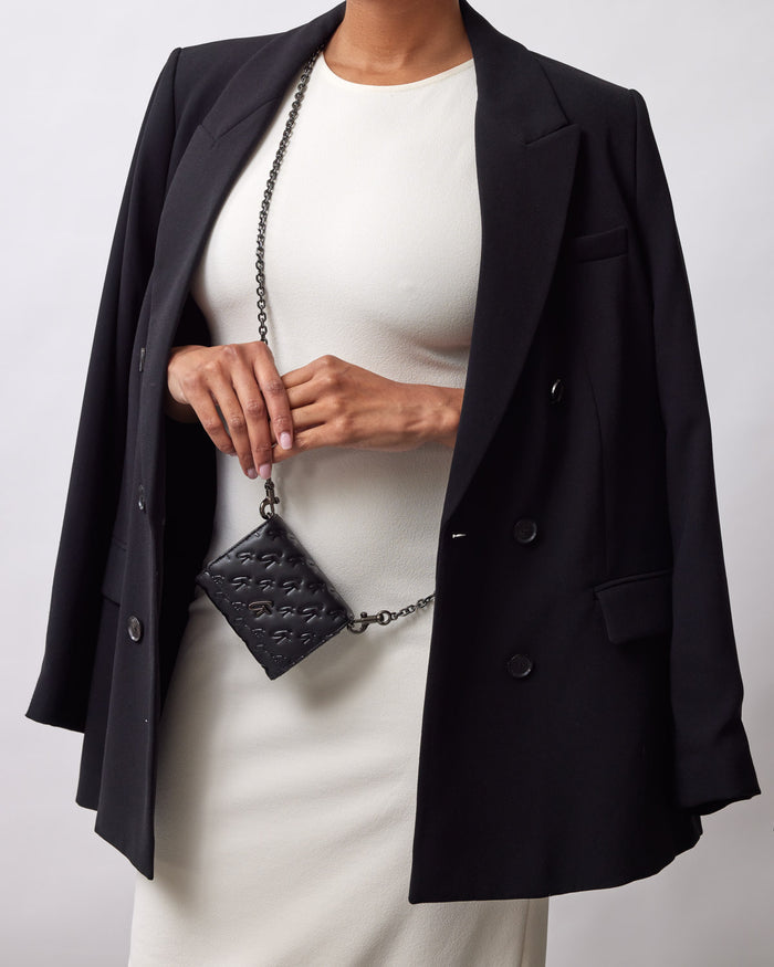 Close-up of a woman holding a black Monogram Gunmetal Compact WOC bag with a chain strap, dressed in a black blazer and white dress.