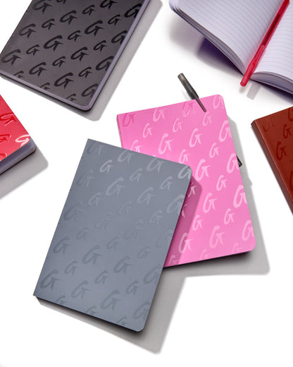 Colorful Monogram notebooks in gray, pink, red, and black with embossed G pattern, displayed on a white surface with a pen.