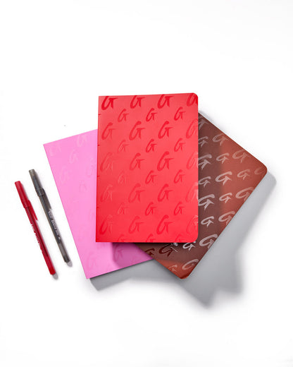 Red monogram notebook with embossed G pattern, accompanied by pink and brown notebooks and red and black pens.