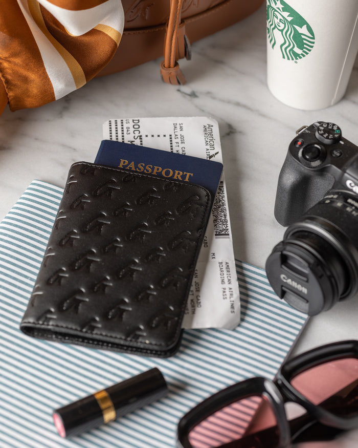 Black monogram passport holder with embossed logo, holding a blue passport, placed on travel documents on a marble surface.