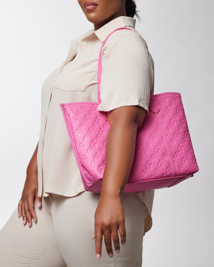 Woman holding a hot pink Monogram Sleek Tote bag with embossed logo pattern, casual beige outfit, against a plain background.