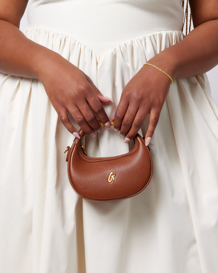 Brown Nano Pebble Hobo Bag with gold logo, held by a person in a white dress, showcasing a compact, textured leather design.