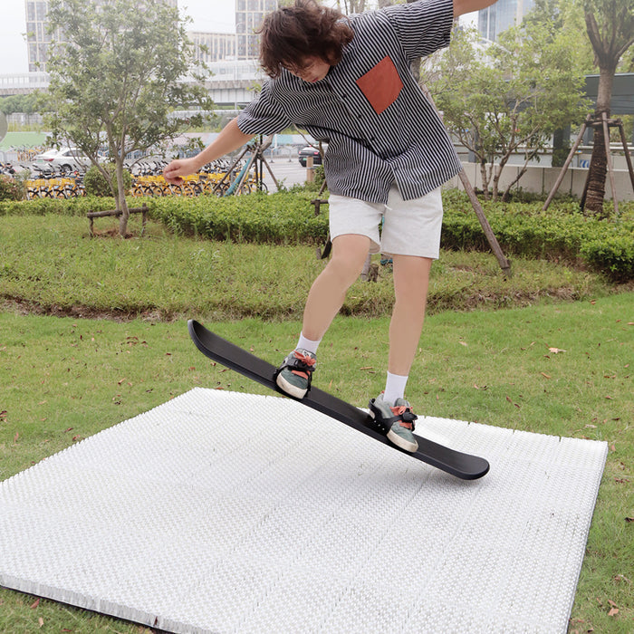 A person on a ski board performing a trick on ski mat in an outdoor setting with greenery and a building in the background.