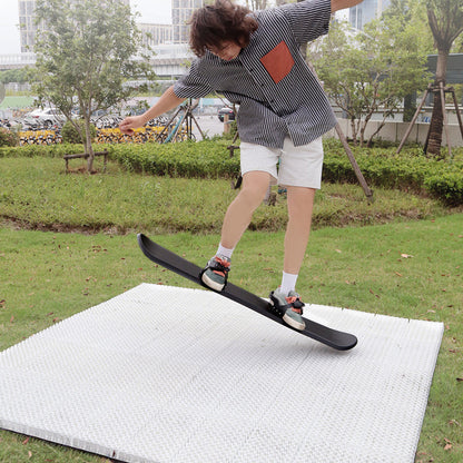 A person on a ski board performing a trick on ski mat in an outdoor setting with greenery and a building in the background.