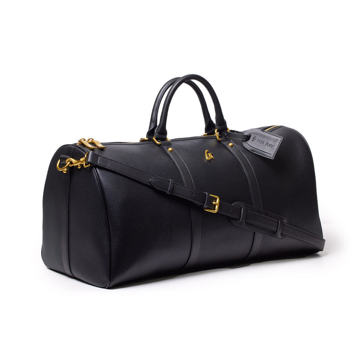 Black Pebble Duffle Bag with gold hardware, dual handles, adjustable shoulder strap, and luggage tag.