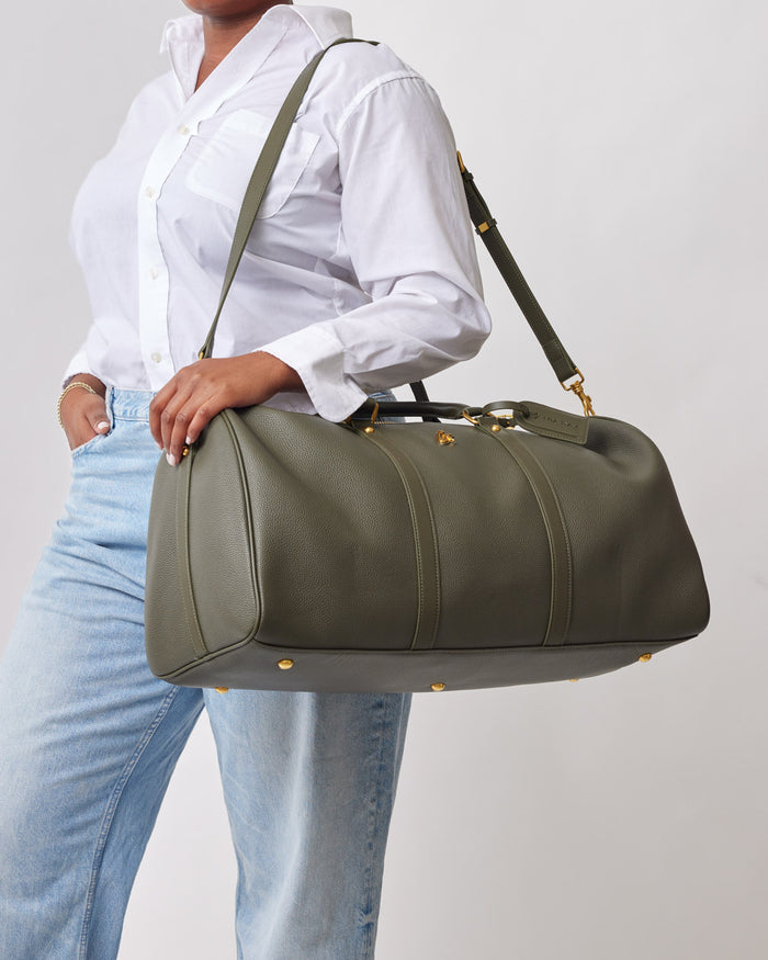 Olive green Pebble Duffle Bag with gold accents, carried by a person in a white shirt and light jeans.
