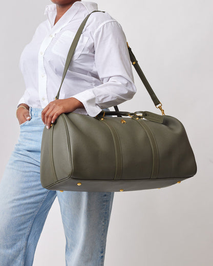Olive green Pebble Duffle Bag with gold accents, carried by a person in a white shirt and light jeans.