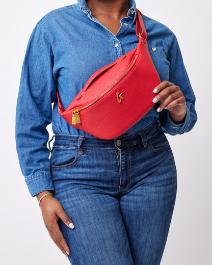 Red pebble-finish fanny pack with gold zipper and logo, worn across the chest of a person in a denim shirt and jeans.