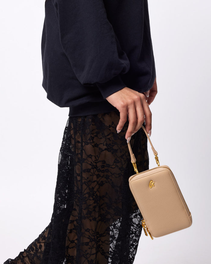A person holding a beige Pebble Phone Crossbody bag with a gold logo, wearing a black sweatshirt and black lace skirt.