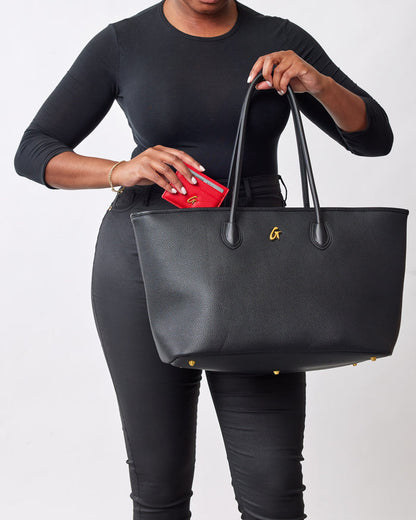 Black pebble tote bag with gold accents, held by a woman in black clothing, with a red wallet partially visible inside.