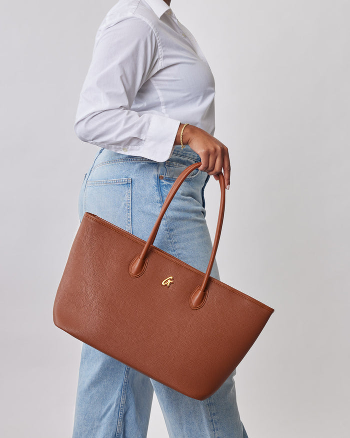 Brown pebble tote bag with dual handles, gold logo, and textured finish, carried by a person in a white shirt and light blue jeans.