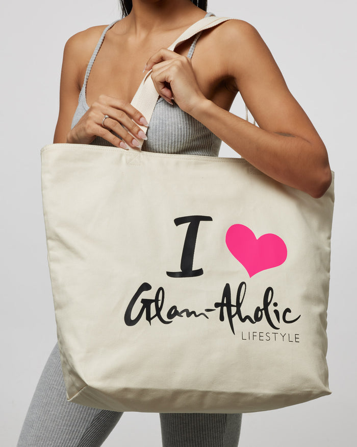 Hot pink logo heart on a cream canvas tote bag with "I Love Glam-Aholic Lifestyle" text, held by a woman in casual attire.
